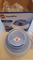 Life brand cool mist humidifier with vented circular top, white and blue plastic body, power cord visible, front view with original box behind.