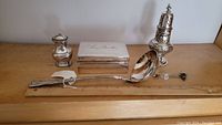 Five silver and silver plate items arranged: a sugar caster, a cigarette box lined with cedar wood, silver shaker, large ladle, and pickle forks.