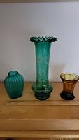 Three glass items on wooden surface with a light background, showing the emerald green vase with enamel decoration, the small blue bottle, and amber glass vase.
