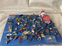 A blue cloth background covered with an assortment of small vintage pins and brooches in enamel and metal, various shapes and themes.