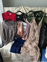 Photo showing a collection of women's clothing on hangers and draped over a couch, including dresses, tops, and pants in various colors and patterns.