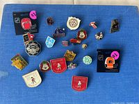 Photo showing an assortment of 23 pins on blue fabric background. Pins vary in design including sports team logos, local badges, and novelty themes. Two pins on black backing cards have $1 stickers on them.