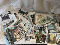 Photo of a large assortment of vintage postcards laid out on a white surface, showing various images including cityscapes, floral designs, people in period clothing, and architectural landmarks.