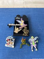 Photo of six Disney themed pins on a blue background, featuring multiple Tinker Bell pins along with Mickey and Minnie Mouse pin with castle and a pin with the text 'It's all about me!'