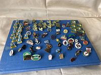 Collection of different travel-themed pins arranged on blue fabric background showing various designs and colors.