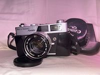 Vintage Bell & Howell Canon QL film camera with Canon 45mm f/1.9 lens, leather carrying case, and lens cap on pink backdrop.