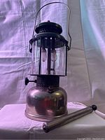 Full view frontal photo of vintage metal Coleman gas lantern with glass globe and attached hanging handle; shows included cylindrical pump accessory in front.