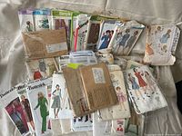 Photo showing a varied collection of vintage paper sewing pattern envelopes, some stacked and some spread out, featuring clothing designs for women and children from brands like Butterick, Simplicity, and McCall's.