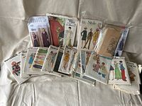 Lot of assorted vintage sewing patterns spread out on a beige fabric surface. The patterns include various brands such as Butterick, Vogue, Simplicity, Kwik Sew, and Style with diverse mid-20th century styles.