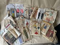 A collection of 15 vintage sewing pattern envelopes laid out on a beige surface, showing multiple brands including McCall's, Simplicity, and New Look with various dress and clothing designs illustrated on them.