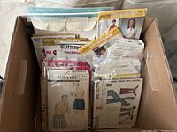 Box of assorted vintage sewing patterns showing multiple branded envelopes like Simplicity, Butterick, Advance, McCall with illustrations of clothes and attachments.