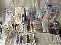 Photo showing a collection of various vintage sewing patterns in plastic bags arranged on a cloth surface featuring mainly women's fashionable dresses, suits, coats, and hats.