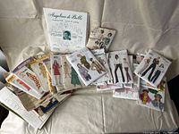 Several vintage sewing patterns spread out on a cream fabric background. Envelopes show illustrations of women modeling various dresses, skirts, jackets, pantsuits and coats. The variety of patterns indicates a mid-20th century fashion theme.