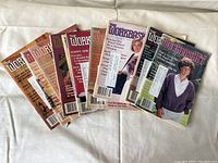 Eight vintage The Workbasket workshop booklets laid out in a slightly curved line on a white fabric background, showing various colorful covers with women modeling handmade clothing and craft project images.