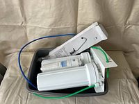 Photo showing two white water filter housings with green and blue tubes attached alongside mounting hardware wrapped in plastic.