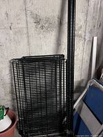 Four stacked black metal wire mesh shelves leaning against a wall in a basement or storage area.