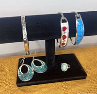 Three silver bracelets displayed on black jewelry stand, showing floral engraving, red hearts inlay, and turquoise inlay designs