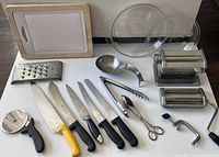 Full view of the lot laid out on the floor showing the cutting boards, knives, ladle, pizza cutter, tongs, cheese grater, glass bowl, manual pasta maker, and scissors.