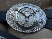 Close-up photo of longhorn steer metal belt buckle showing embossed design with laurel wreath and floral border.