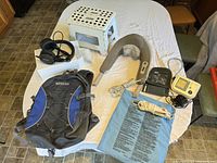Photo showing full assortment of household items on table: folding foot stool, black and blue backpack, TV headphones, neck massager, heating pad, blood pressure monitor with charger and cuff.