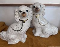 Photo showing matching pair of white porcelain dog figures with crackle glaze and gold accents, sitting side by side on a brown textured surface.