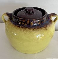Front view of yellow bean pot with brown and blue rim glaze and dark brown lid with knob showing the shiny glazed surface and two handles on the pot.