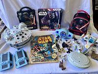 Full assortment of Star Wars collectibles including Millennium Falcon, trivia game, comic, ships, figures, and lunch bag lined up on white backdrop.