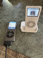 Two iPods side by side on table, one silver iPod Nano showing menus, one white iPod Classic in dock showing album art.