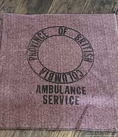Close-up of part of the wool blanket showing 'Province of British Columbia Ambulance Service' stamped in black on red and white herringbone wool fabric.