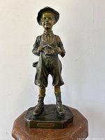 Front view of the bronzed metal statue "Le Gamin" showing the standing boy with facial features, hat, suspenders, and boots on base with nameplate.