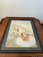 Framed art print of a nude woman reclining on sand, viewed from above. Dark wooden frame with black matte border.