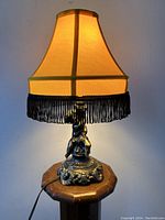Front view of the lit lamp on wooden surface showing bronzed metal cherub figurine base and fringed fabric lampshade.