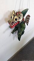 Left angled view of the dragon puppet showing front and side of the detailed carved and painted face, limbs, and wings suspended by strings.