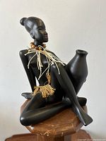 Front view showing the seated Nubian lady holding the vase, wearing a bead necklace and raffia skirt, placed on a wooden stool.