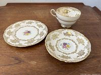 Set of three tea cup, saucer, and side plate shown individually on a wooden surface, highlighting the gilt gold detailing and floral center.