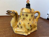 Side view of the camel-shaped porcelain teapot with rider figurine on lid showing body, handle and spout.