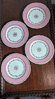 Four antique English porcelain dinner plates with hot pink border, gold trim, and floral pattern displayed on a dark wooden surface.