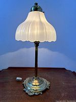 Full view of the vintage table lamp with brass base and opaque white glass bellflower shade on a wooden surface against a blue background. Lamp is switched on, showing the illumination through the shade.