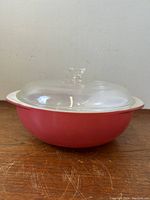 Front view of vintage Pyrex deep raspberry casserole dish with clear glass lid on wooden surface.