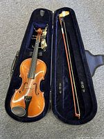 Violin and bow inside open black case with dark blue plush lining, showing violin body and bow side-by-side
