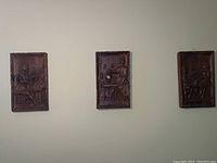 Three carved wood rectangular plaques mounted on a wall, each showing a seated person in detailed carving.
