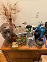 Wide view of lot showing various metal art, pottery, glass spray bottles, wooden bird, and dried wheat stalks on wooden cabinet.