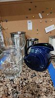 Four pitchers including cobalt blue ceramic, clear glass, and two stainless steel, plus clear glass vase on granite countertop with corkboard background.