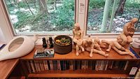 Full view of lot on wooden shelf including sculptures of human figures, ceramic vintage bed pan, artificial succulent in pottery, small antique binoculars and patterned black and white glass art piece.