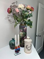 Lot contents on white surface showing tall floral bud vase with faux flowers, green pottery vase, colorful block candlestick with candles, and white ceramic covered jar