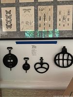 Four vintage cast iron tools displayed on wall beneath framed insect and butterfly prints. Items include fireplace stampers and a cattle water insert level, showing overall condition and design details.