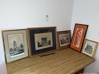Five framed wall art pieces shown on wooden surface against white wall, including black and white etchings and one red figurative piece.