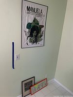 Framed poster titled 'MANUELA' hanging on light green wall, with two smaller framed pieces below on floor