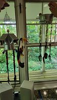 Photo showing three distinct wind chimes hanging in front of a window with outdoor greenery background: metal birdhouse style, colorful bead and metal tubes, and toucan shaped chime.