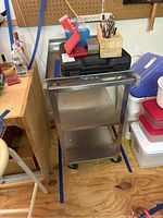 Full view of the metal work cart showing three tiers with some items on the top shelf (items not included in the lot). The cart has a push handle and is on casters.